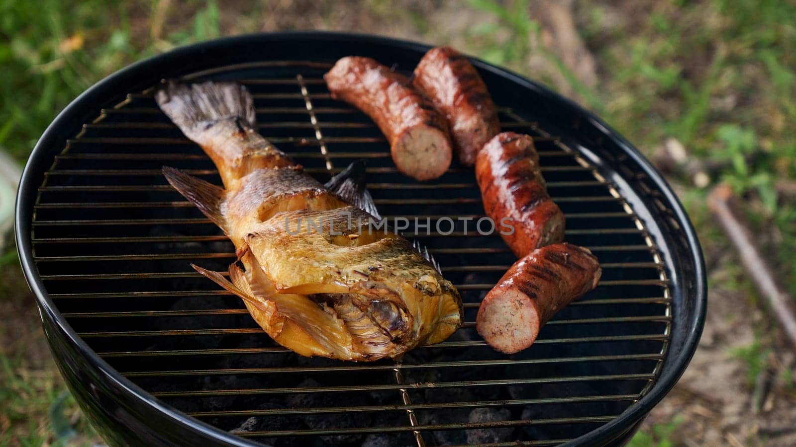 Top view of fresh grilled whole fish, skewer, sausages on round black charcoal grill, green grass background. Barbecue, grill and food concept. Camping with family