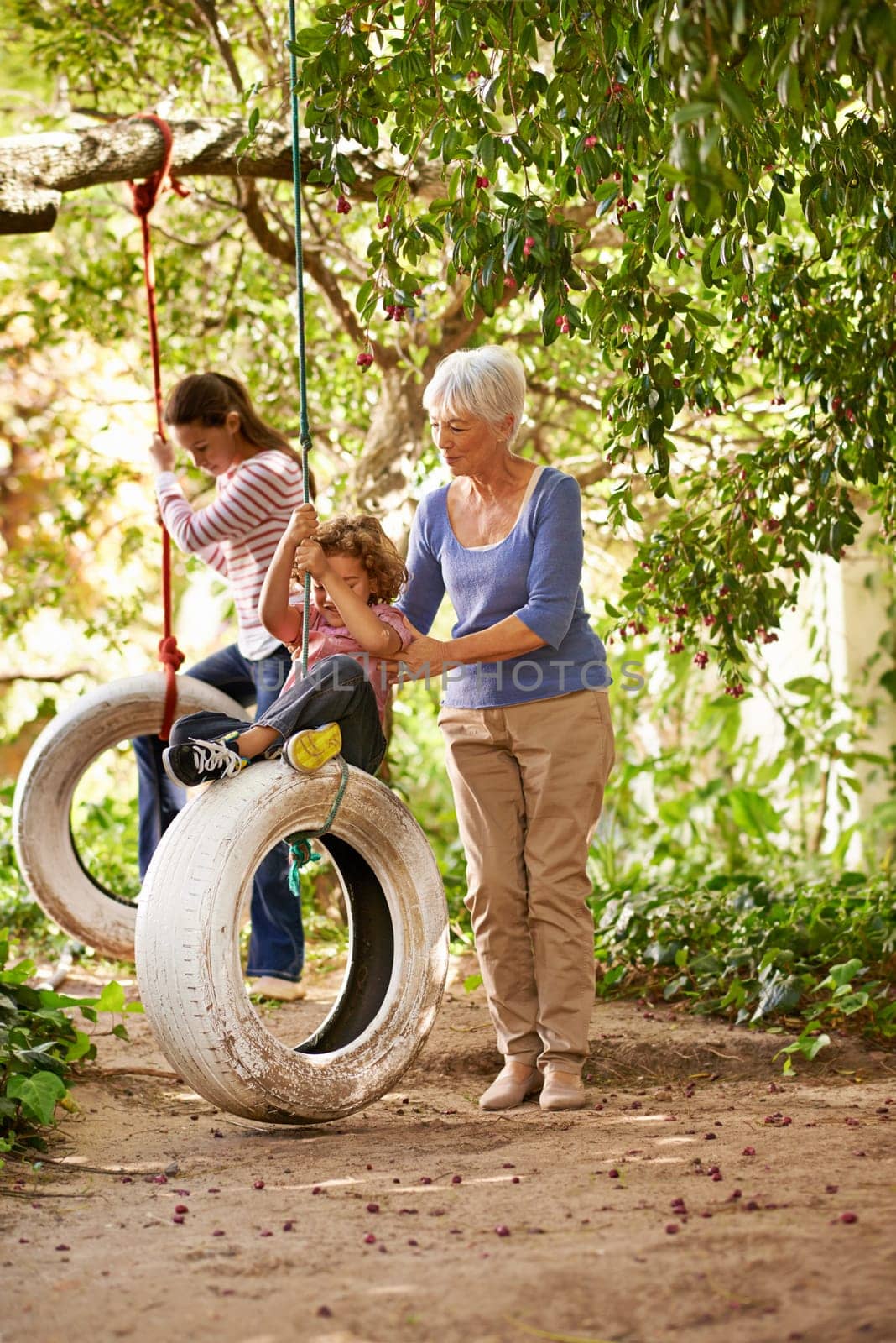 Senior woman, garden and grandma pushing her grandchildren on a tyre swing or holidays and having fun in summer. Excited, grandkids and elderly woman outdoors on jungle gym or on sunny day at a park.