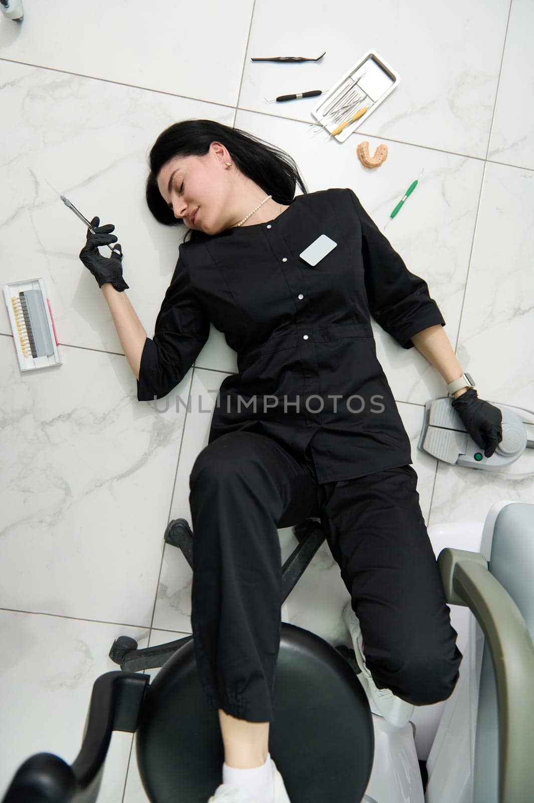 Conceptual portrait of a young hardworking female dentist hygienist, orthodontist surgeon, holding a syringe and lying on the floor in a dental office, sleeping, feeling tired after a hard day at work
