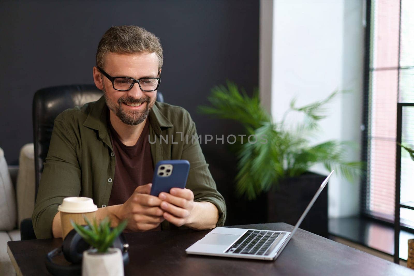 Positive freelancer who working from comfort of home. Smiling man seen texting message on phone while taking well-deserved break from work, resting near notebook. . High quality photo
