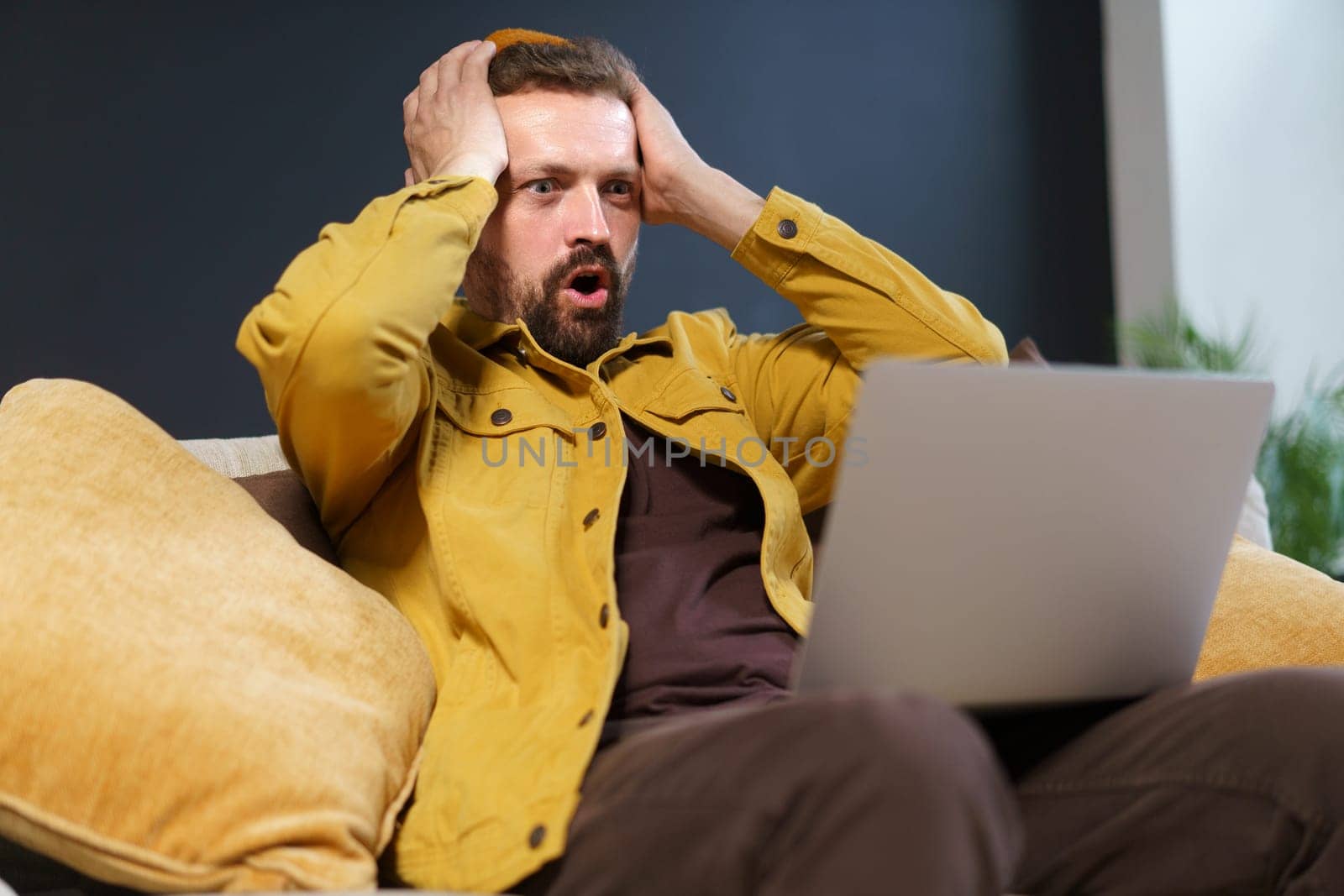 Shocking discovery made by internet user. Man holding head with two hands, expressing strong sense of shock and disbelief as he views content displayed on screen of laptop. by LipikStockMedia