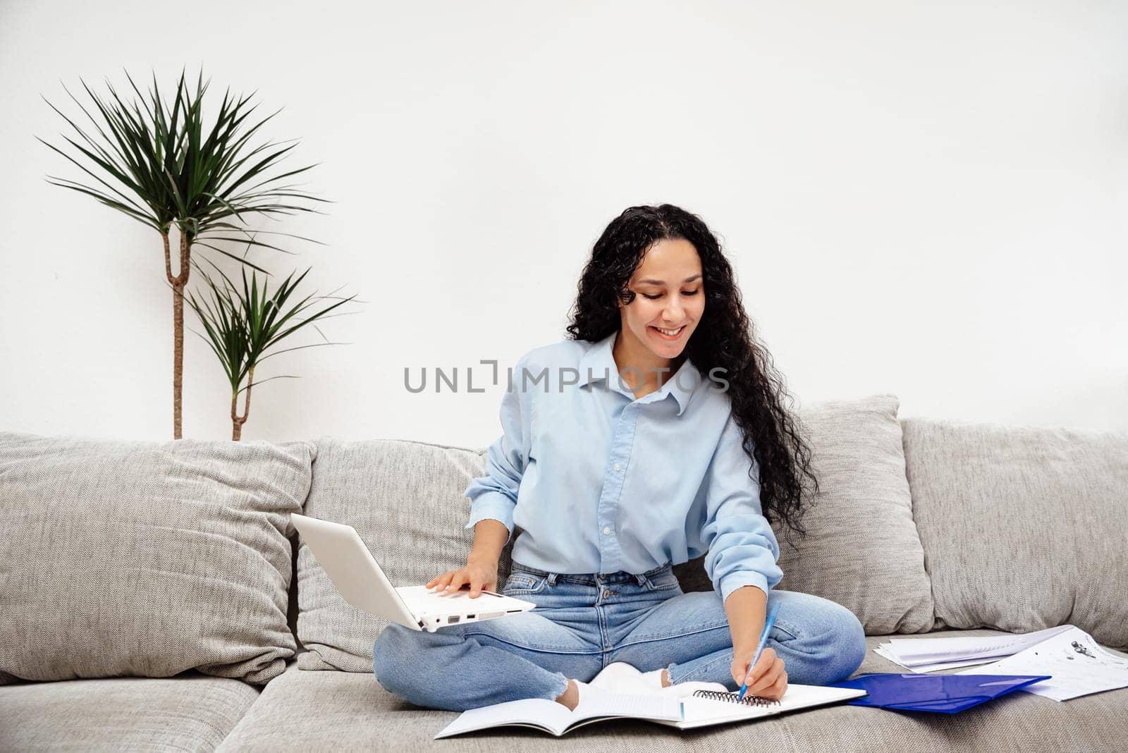 Beautiful young Arab woman sits on the sofa in the workspace and writes down homework. One hand on a laptop the other is taking notes