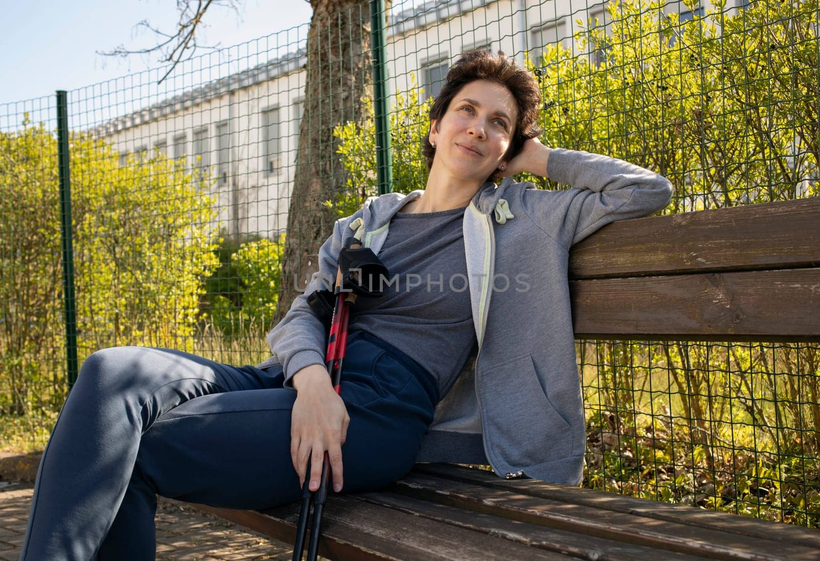 Happy Resting, Relaxing Woman Sits on Bench Holding Poles After Nordic Walking. Beautiful Happy Caucasian Woman Enjoys Sunny Day. Physical Activity With Walking Poles. Outdoor Recreation. Horizontal.