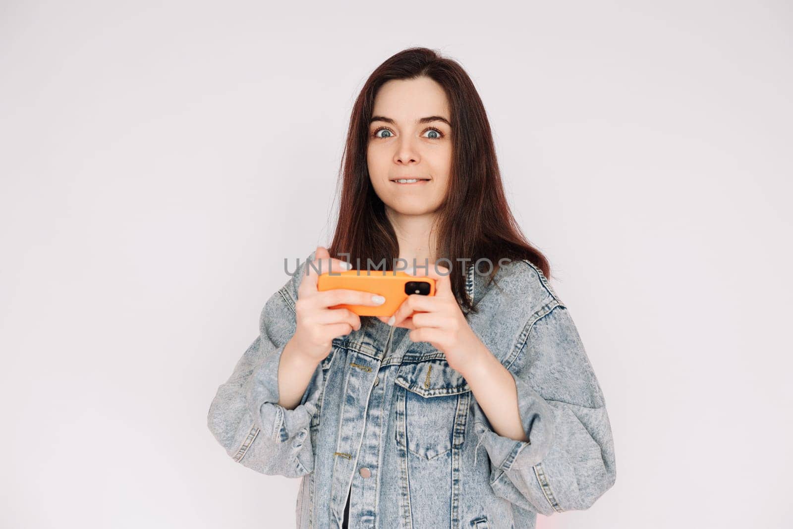 Indecisive and Insecure: Young Woman in Denim Jacket Playing Smartphone Game, Isolated on Gray Background. Emphasizing Uncertainty and Doubt.