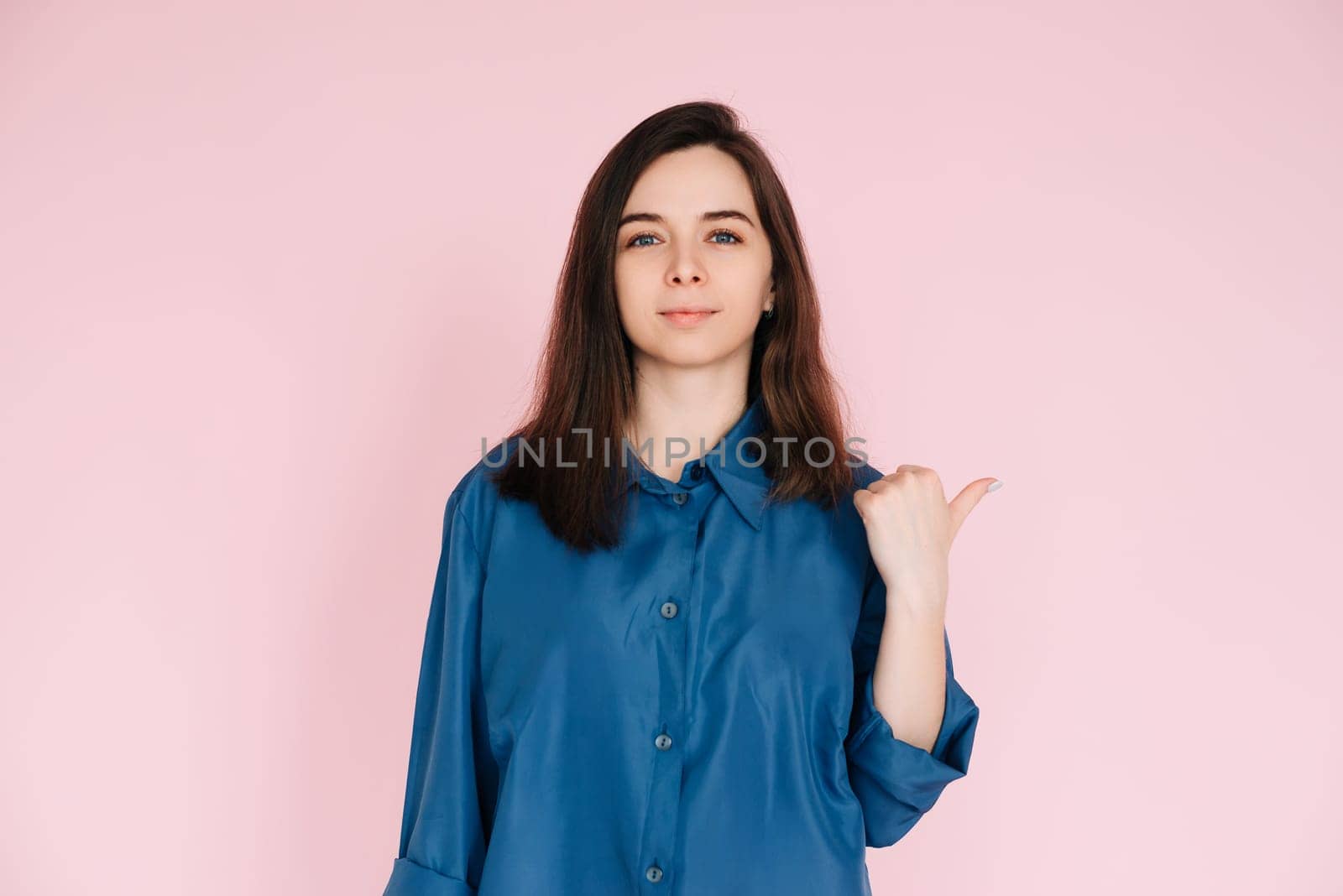Young Businesswoman in Blue Shirt Pointing at Mockup, Symbolizing Office Promotion and New Opportunities: Professional Portrait on Pink Background.