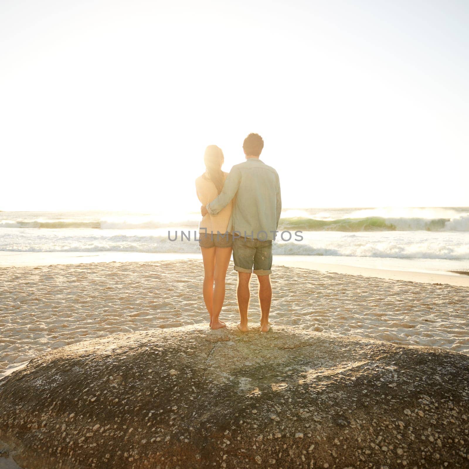 Sweethearts at sunset. Rearview shot of a young couple enjoying the sunset at the beach. by YuriArcurs