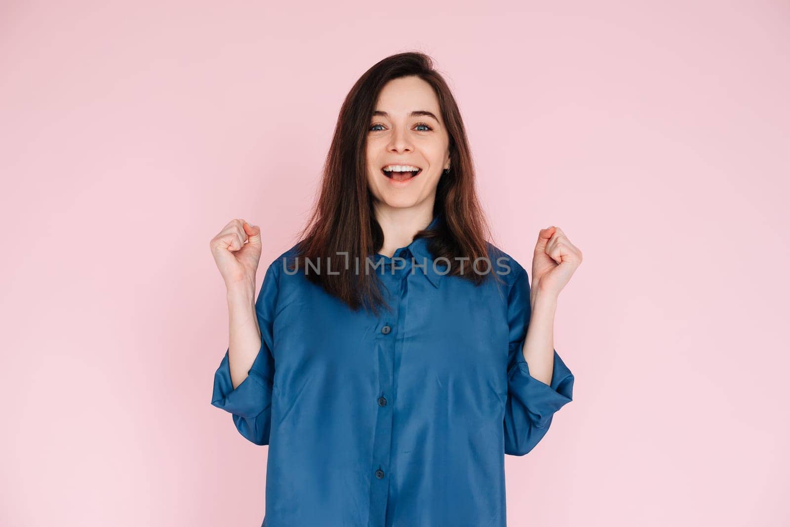 Euphoric Woman Celebrating Success, Raising Fists in Triumph: Isolated Pink Background Portrait. Joyful Woman Celebrating Achievement, Raising Fists in Excitement: Isolated Pink Background Portrait.