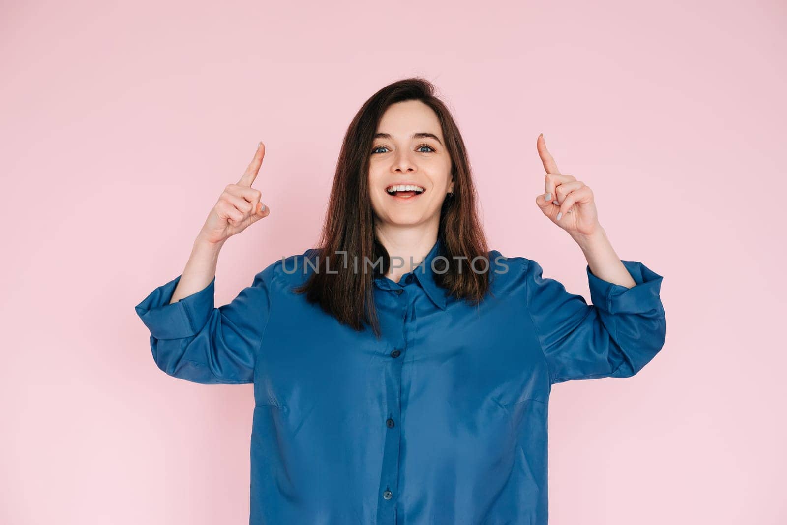 Vibrant Portrait of Energetic Young Woman Expressing Amazement with Open Mouth and Pointing Fingers Upward, Isolated on Pink Background.