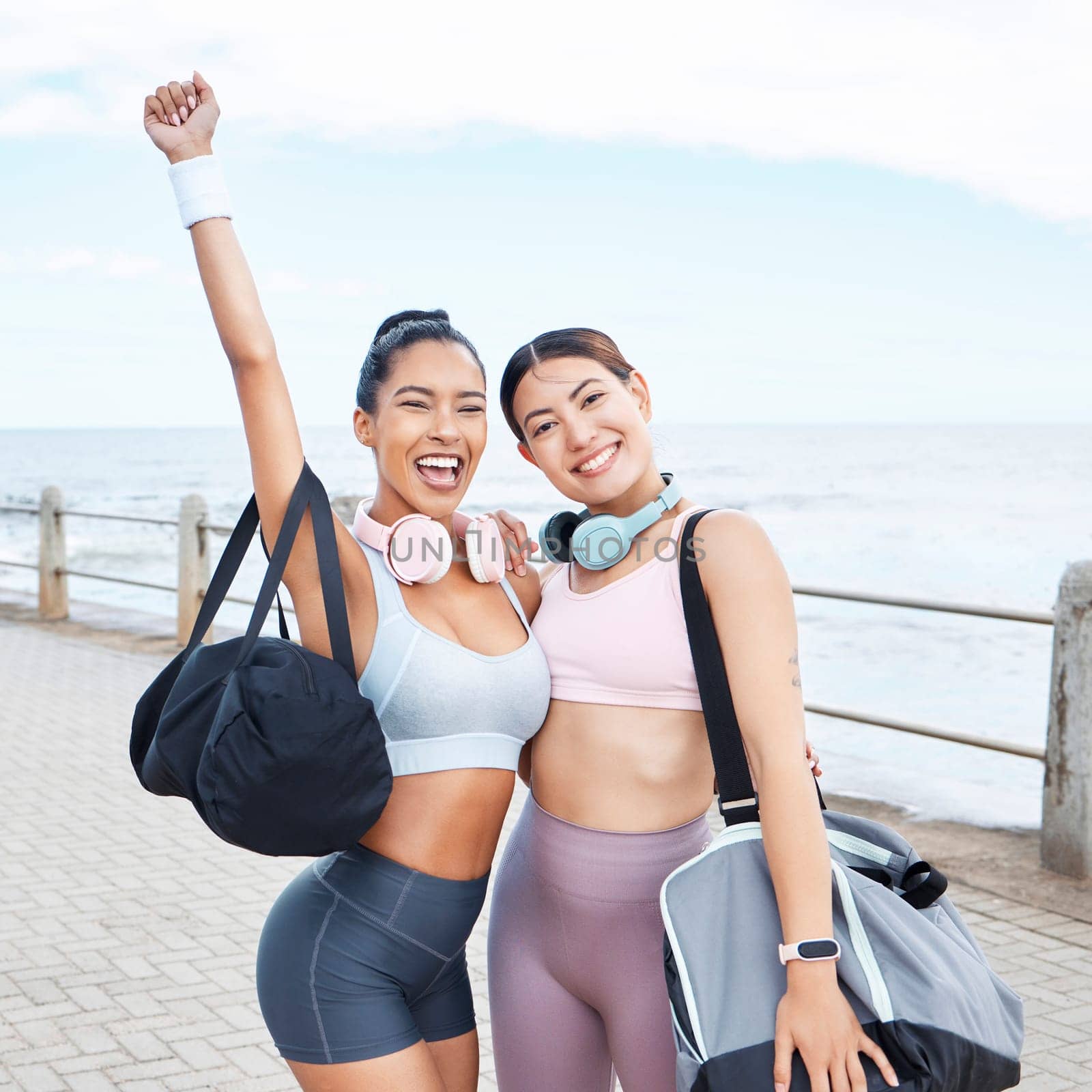 Excited, fitness success and friends exercise by ocean for outdoor training wellness, accountability and happy with body results. Happy sports women with workout bag and headphones for motivation.