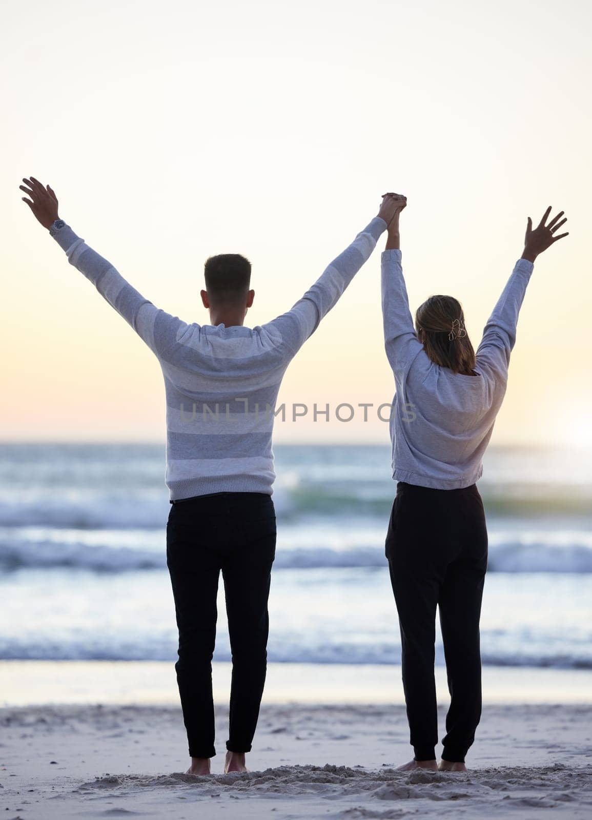 Couple, beach and hands up outdoor while happy at sunset for love, freedom and peace with calm ocean. Young man and woman together on vacation holiday at sea to relax, travel and connect in nature.
