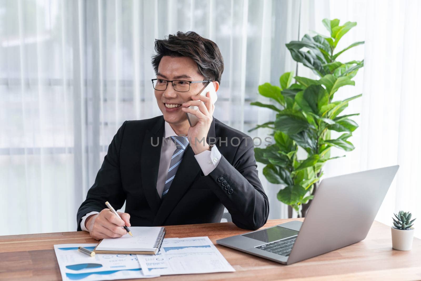 Businessman working on laptop for on office desk workspace with his smartphone. Smart executive researching financial business data or planning strategic business, talking on phone. Jubilant