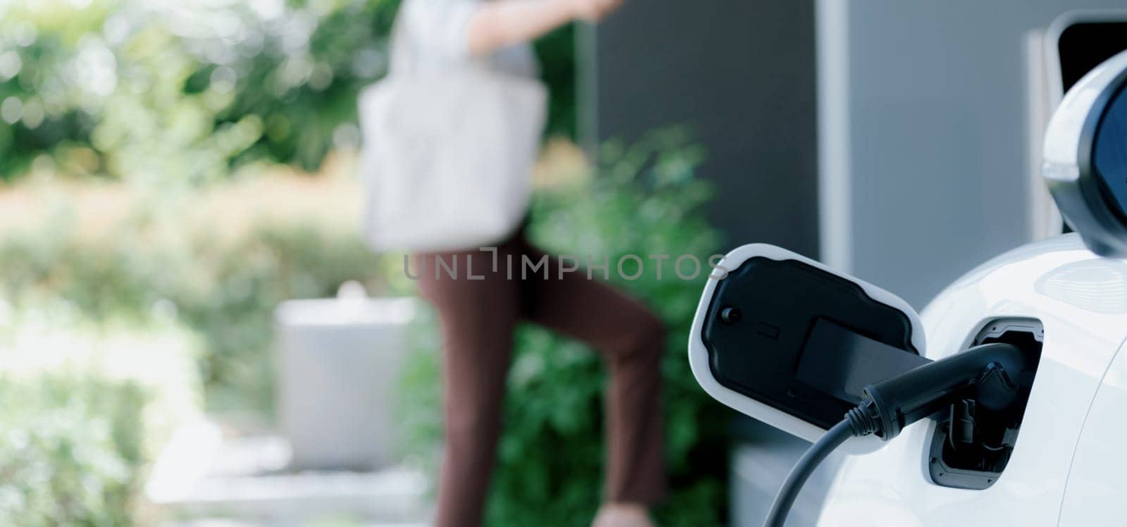 Focus charger plug into EV car with blur backdrop of progressive woman walking. by biancoblue