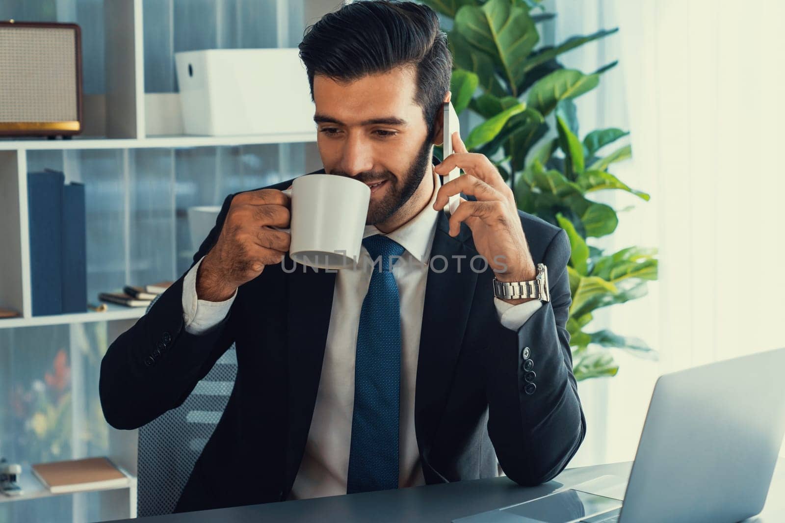 Businessman working in modern office workspace with cup of coffee in his hand while answering phone call making sales calls or managing employee. Fervent