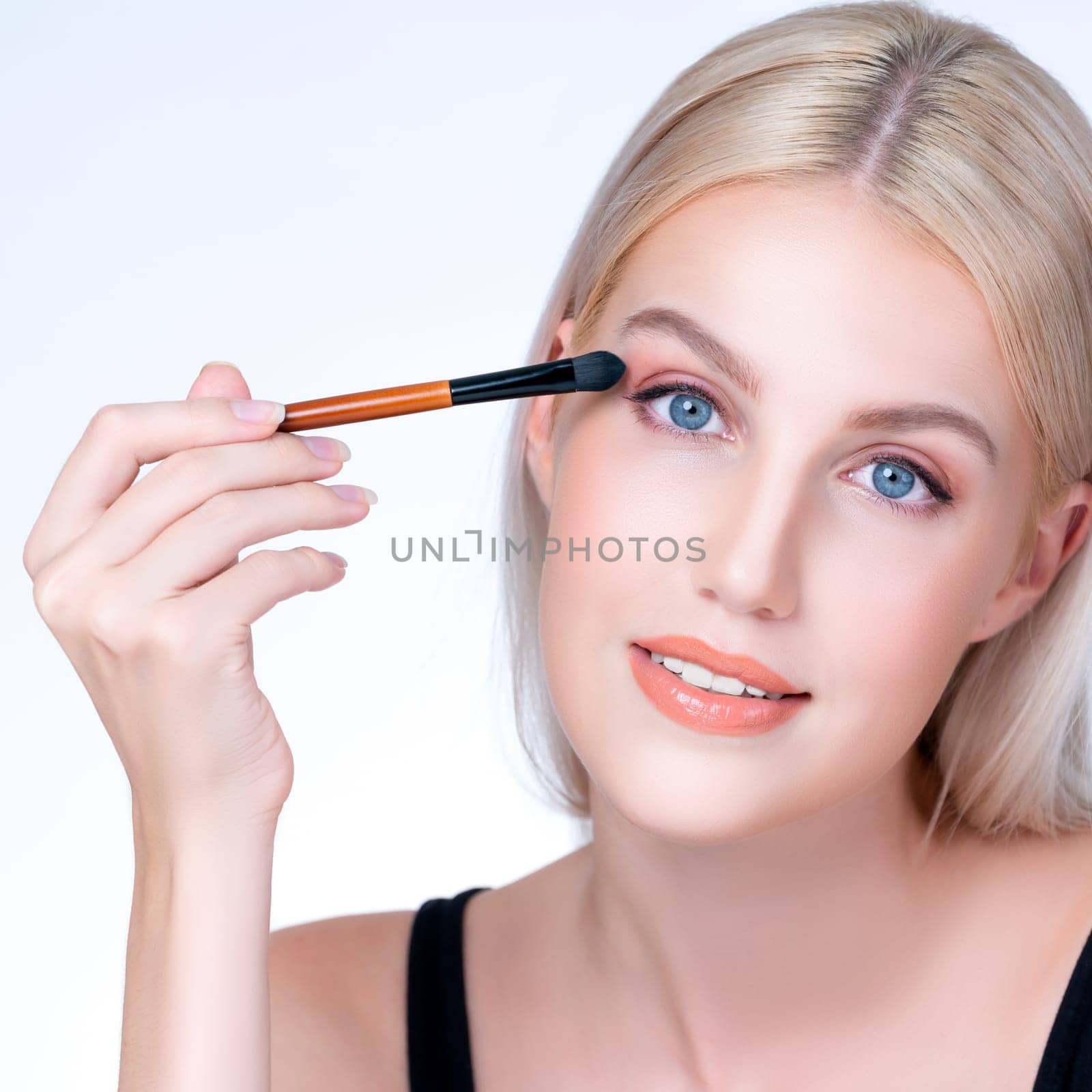 Closeup beautiful personable girl with flawless applying eye shadow makeup with eyeliner brush. Cosmetic facial painting process on lovely young woman with perfect clean skin in isolated background.
