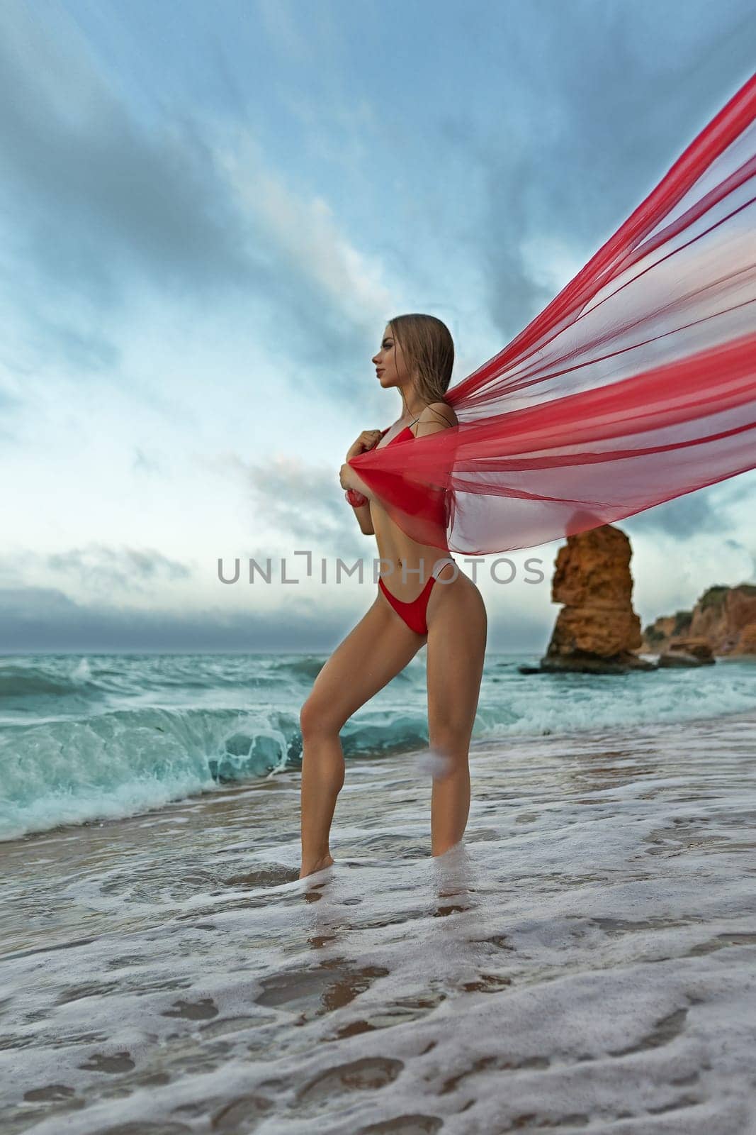A sexy fitness model girl in a red swimsuit stands in the sea, wrapped in a transparent red cloth