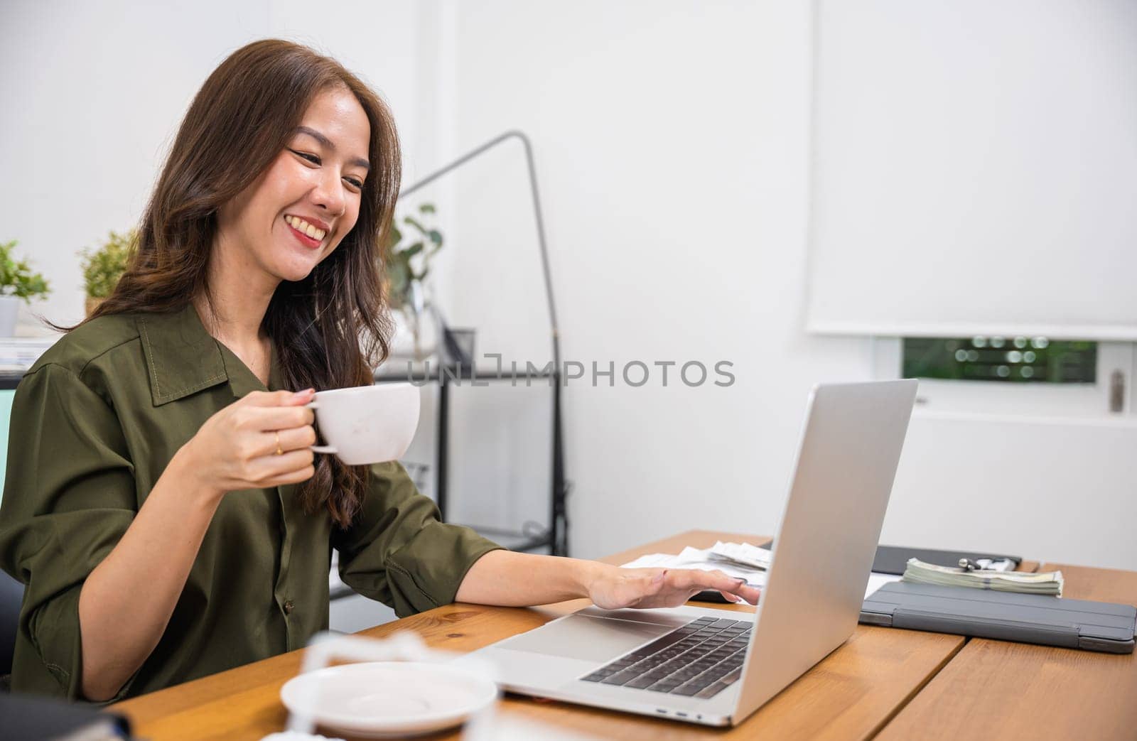 Portrait of beautiful woman holding coffee mug on hand while online shopping for cyber monday sales on laptop computer, smiling young business woman drinks coffee sitting desk using laptop keyboard