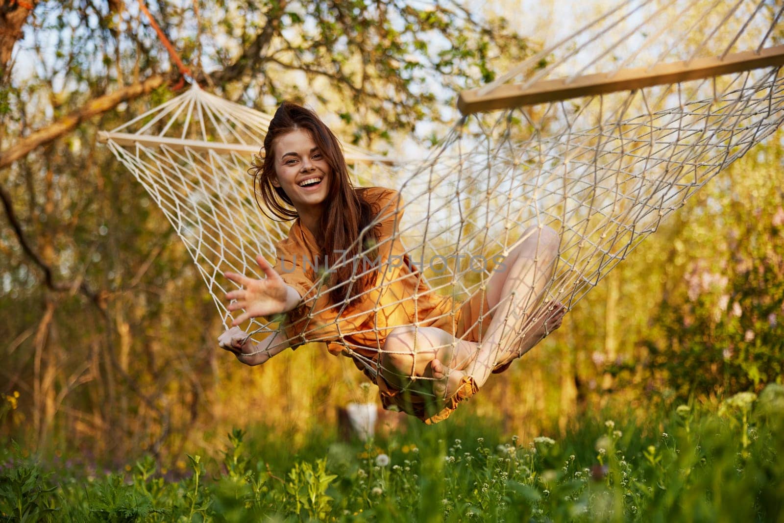 a joyful woman with long hair is lying in a hammock in an orange dress and happily smiling at the camera. A photo taken on the street on the theme of recreation in the country. High quality photo