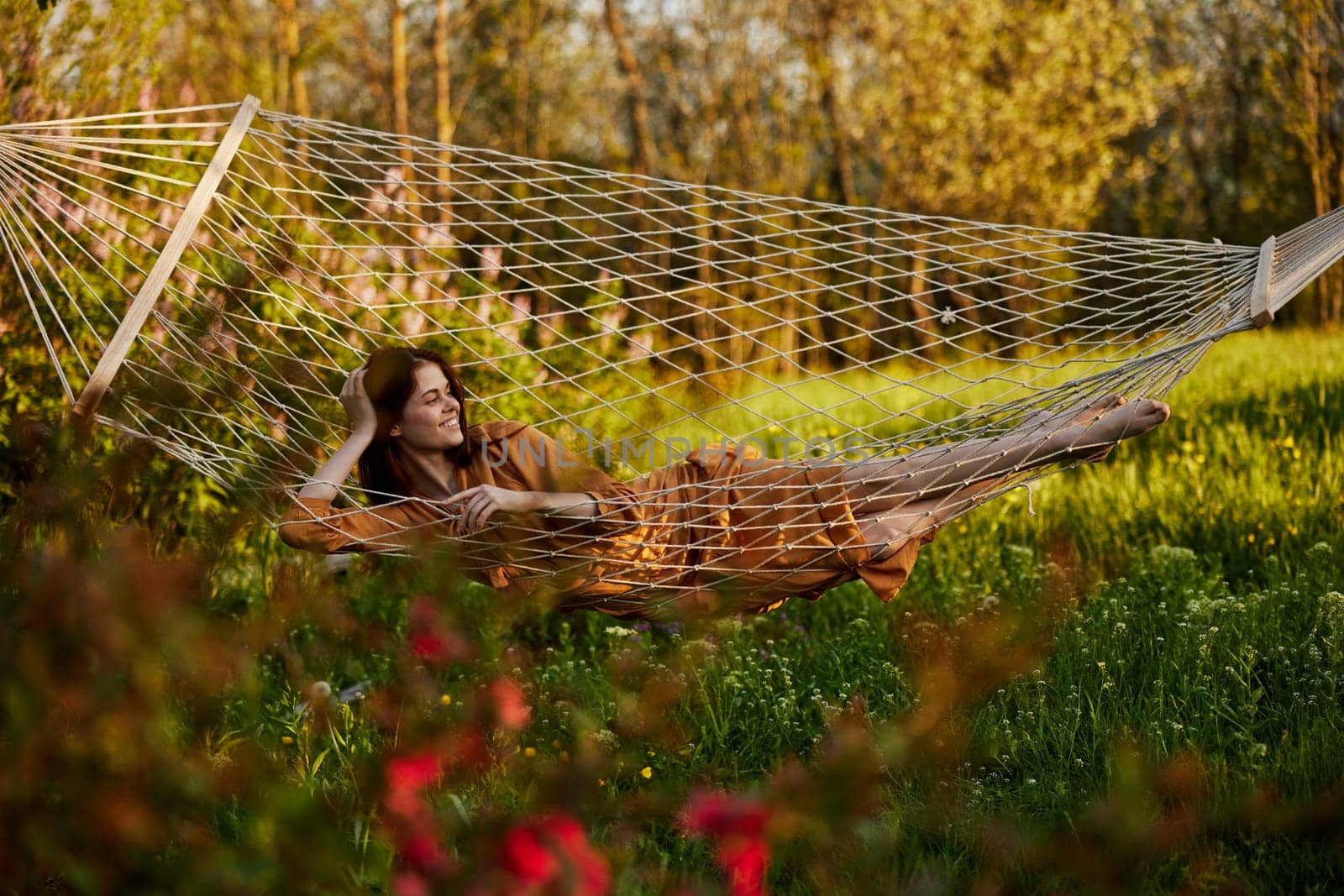 a beautiful woman is resting in nature lying in a mesh hammock in a long orange dress looking to the side, propping her head with her hand. Horizontal photo on the theme of recreation. High quality photo