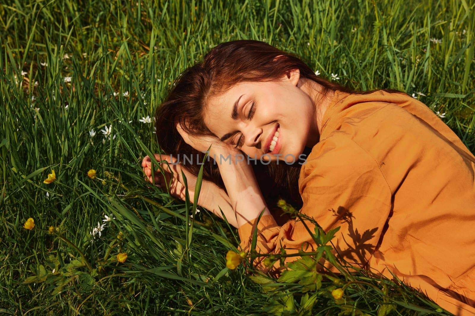 a calm woman with long red hair lies in a green field with yellow flowers, in an orange dress smiling pleasantly, closing her eyes from the bright summer sun by Vichizh