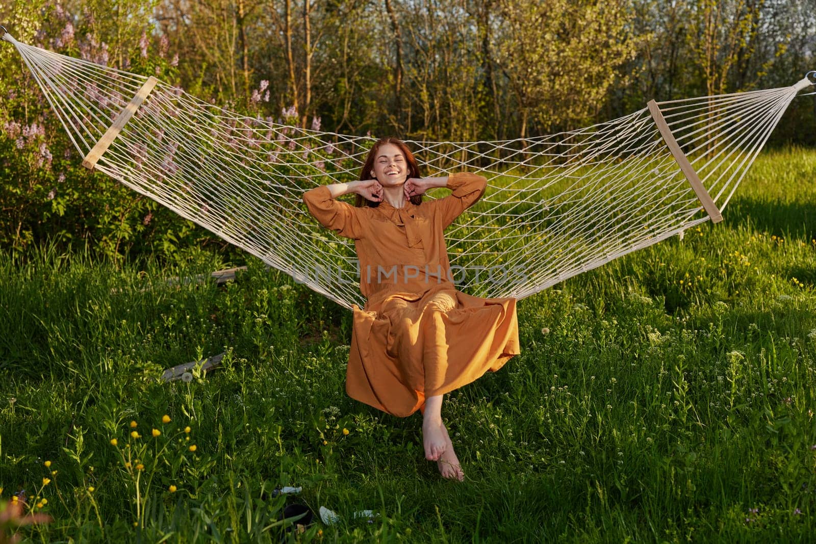 horizontal photo of a beautiful, red-haired woman lying in a hammock enjoying a rest in a long orange dress, on a warm summer day, touching her hair with her hand by Vichizh