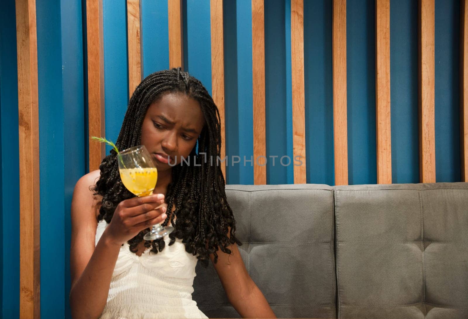 copyspace of afro girl on a gray sofa looking with a look between puzzled and dislike or disgust at her cocktail. High quality photo