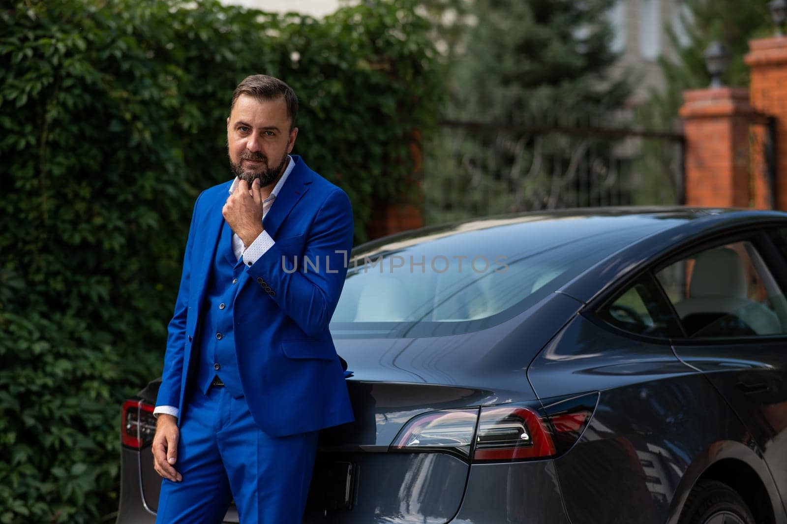 Caucasian bearded man in a blue suit stands near a black car in the countryside in summer