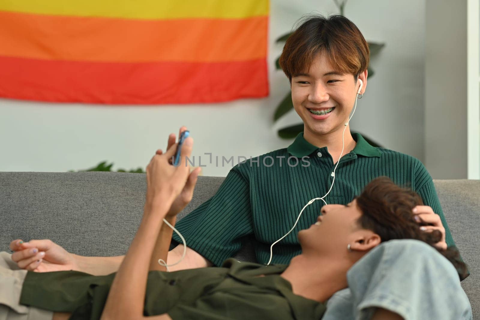 Carefree male gay couple listening to music on earphones, spending time together at home. LGBT, relationship and comfort living concept.