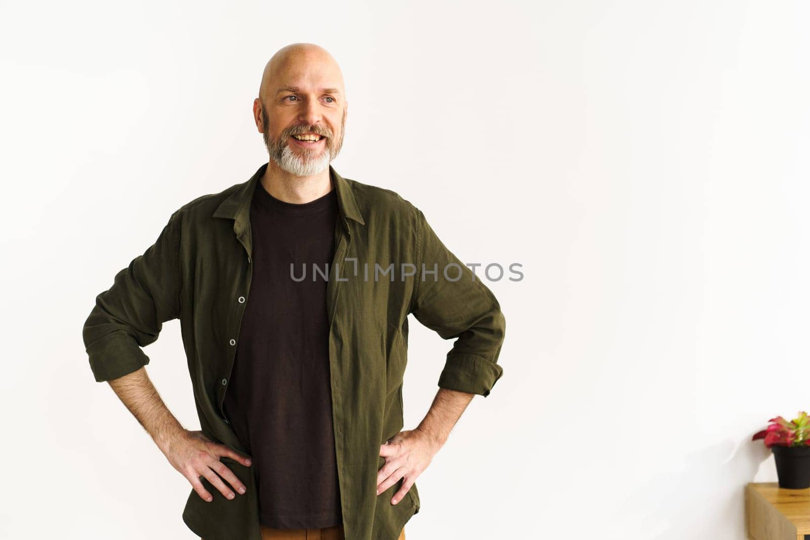 Smiling mid-aged bald man with silver beard standing confidently in front of white wall. His positive posture and expression radiate happiness and joy, showcasing contentment and cheerful demeanor. . High quality photo