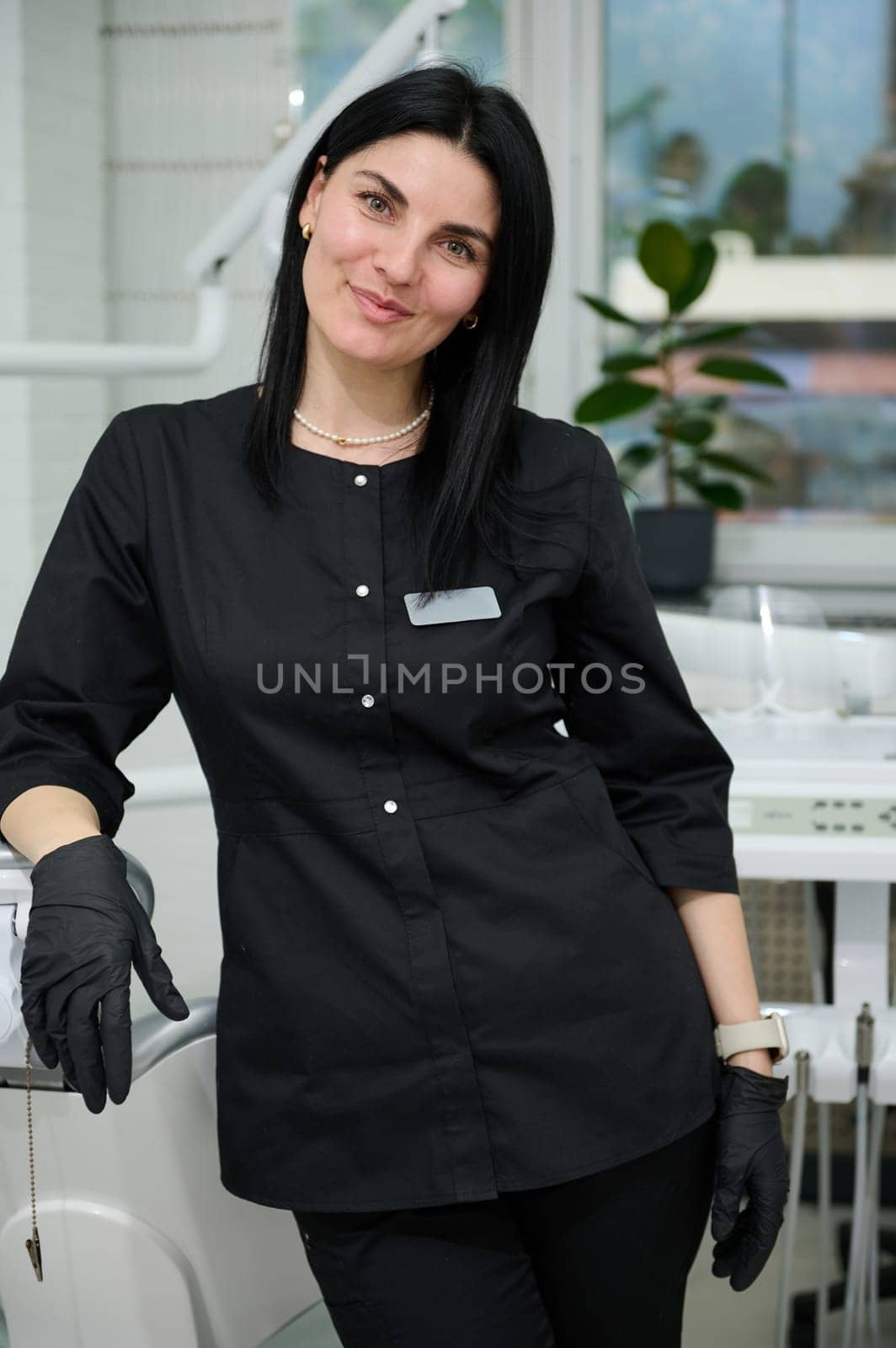Confident portrait of a beautiful experienced woman doctor, orthodontist, dental hygienist wearing stylish black medical unifrom and gloves, smiles looking at camera, standing in newest dentist office