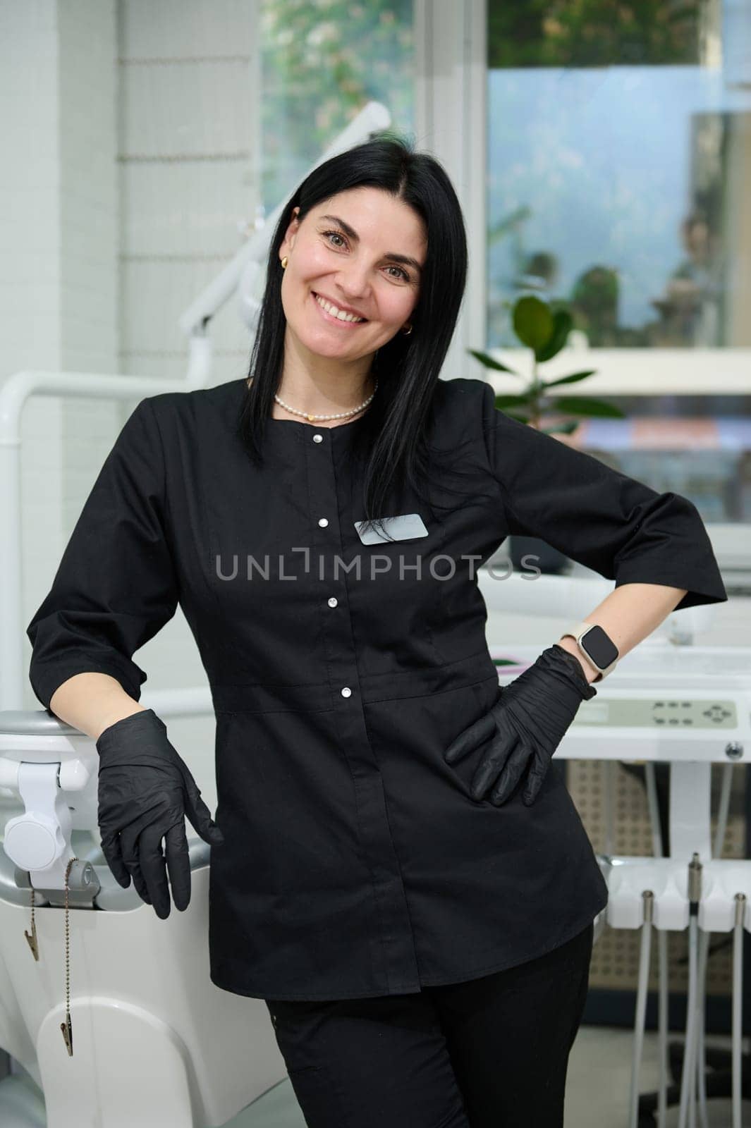 Professional portrait of beautiful middle aged confident female dental hygienist in black uniform, doctor dentist orthodontist, smiling looking at camera, standing by dentist chair in dentistry office
