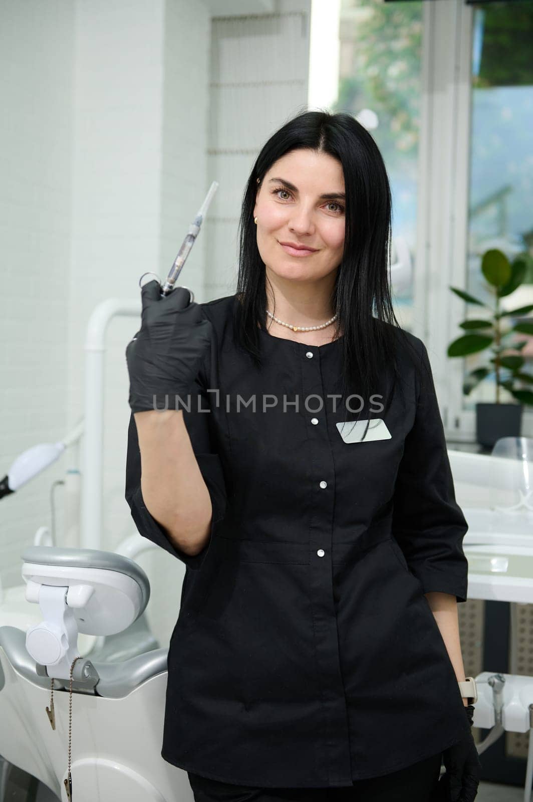 Beautiful confident female dental hygienist, surgeon orthodontist in black medical uniform, holding a syringe with dental anesthesia, looking at camera, standing by dentist chair in dentistry clinic