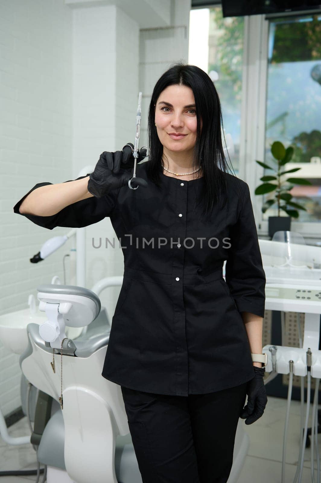 Caucasian young confident professional woman dentist doctor, dressed in stylish black medical uniform, standing by dentist's chair in the dentistry office, holding a syringe with dental anesthetic