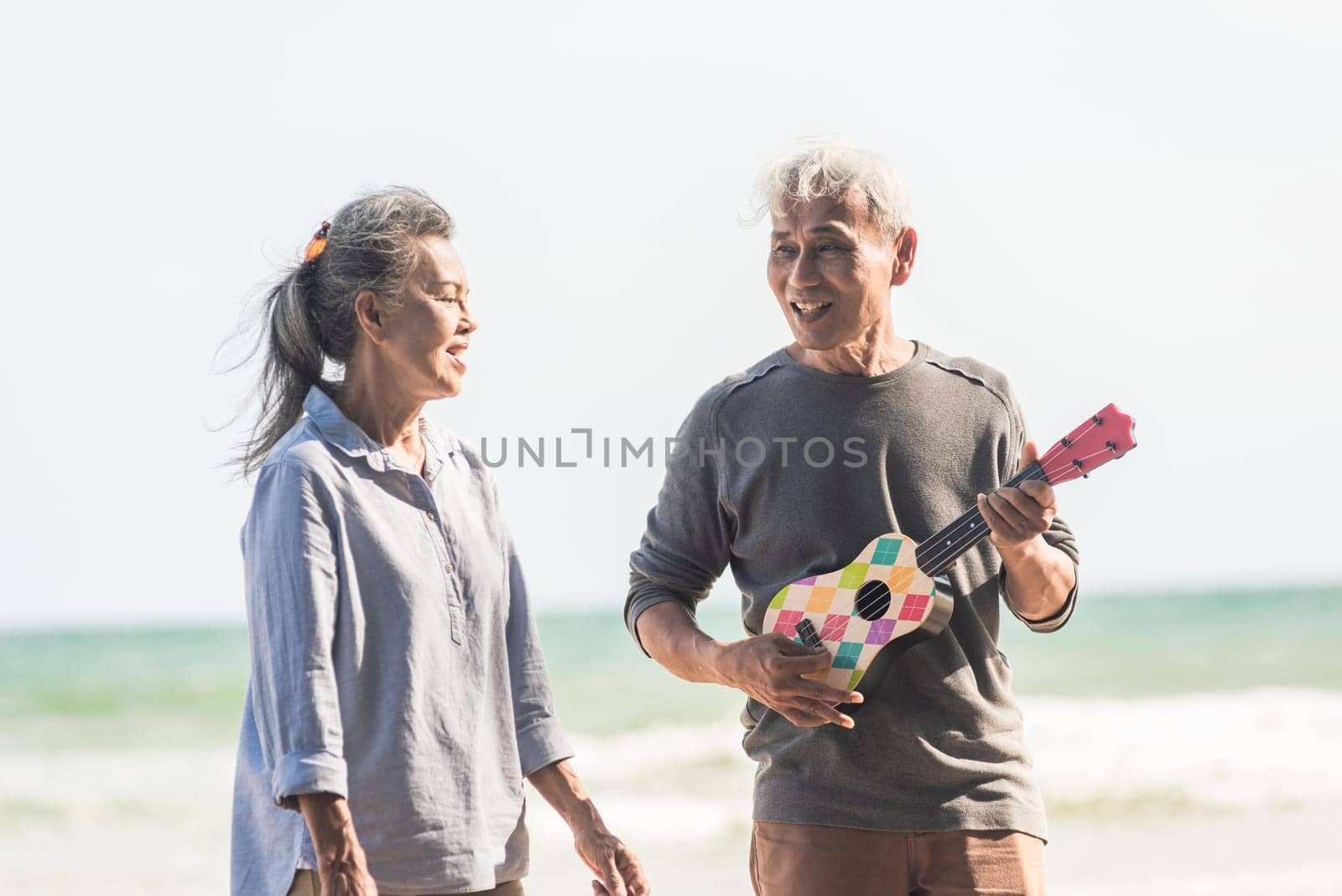 Happy senior couple relaxing outdoors singing and playing acoustic guitar at beach near sea sunny day, Mature man playing ukulele for his wife at sea, plan life insurance at retirement couple concept