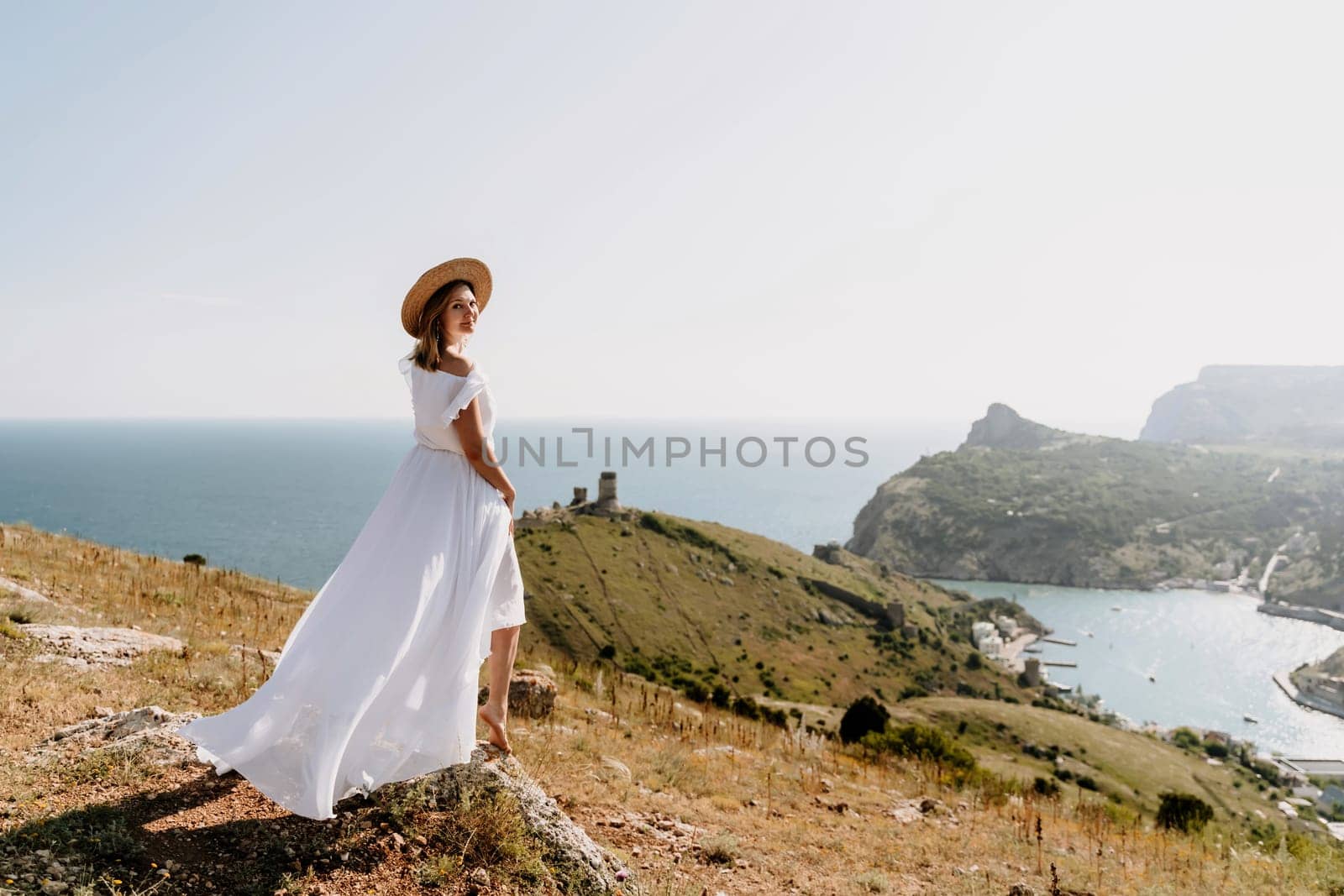 Happy woman in a white dress and hat stands on a rocky cliff above the sea, with the beautiful silhouette of hills in thick fog in the background