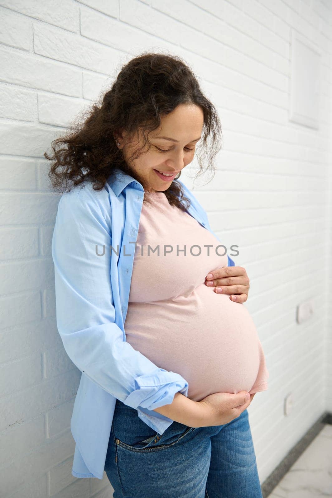 Lovely pregnant woman in 30 week of pregnancy, smiling and gentry stroking her belly, over white brick wall background. by artgf