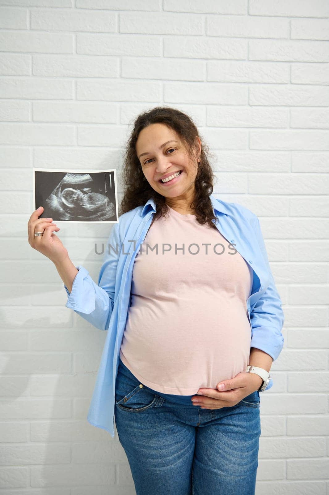Happy African American positive adult pregnant woman, caressing her big belly, smiling and showing at camera the ultrasound of her newborn baby, isolated over white brick wall background. Pregnancy