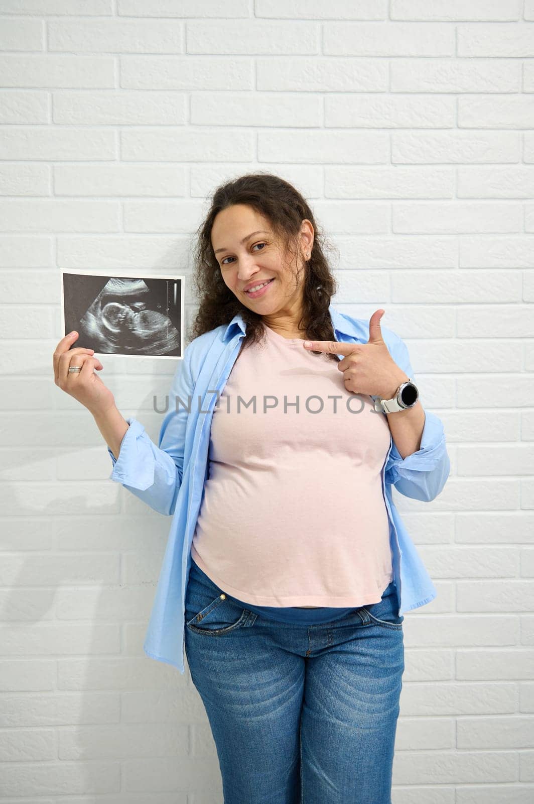 Pregnant woman smiles at the camera and points towards the ultrasound at her baby, isolated against a white brick wall. by artgf