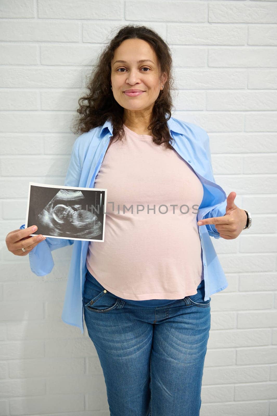 Mature happy pregnant woman pointing at her belly, holding the ultrasonography of her baby, smiling looking at camera by artgf