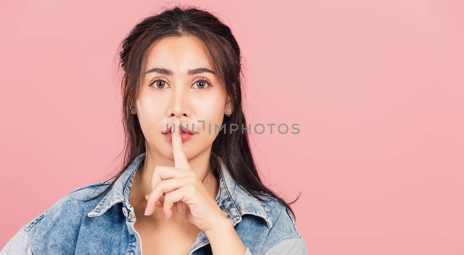 Portrait Asian beautiful young woman wear denim smile stand making finger on lips mouth silent quiet gesture, studio shot isolated on pink background, Thai female quiet hand sign copy space