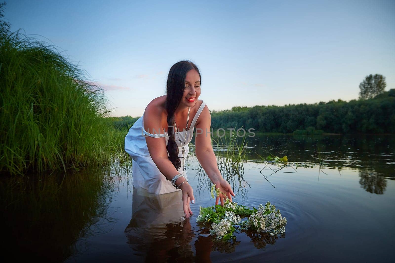 Adult mature brunette woman in a white dress, sundress and a wreath of flowers in summer in water of river or lake in the evening at sunset. Celebration of the Slavic pagan holiday of Ivan Kupala by keleny