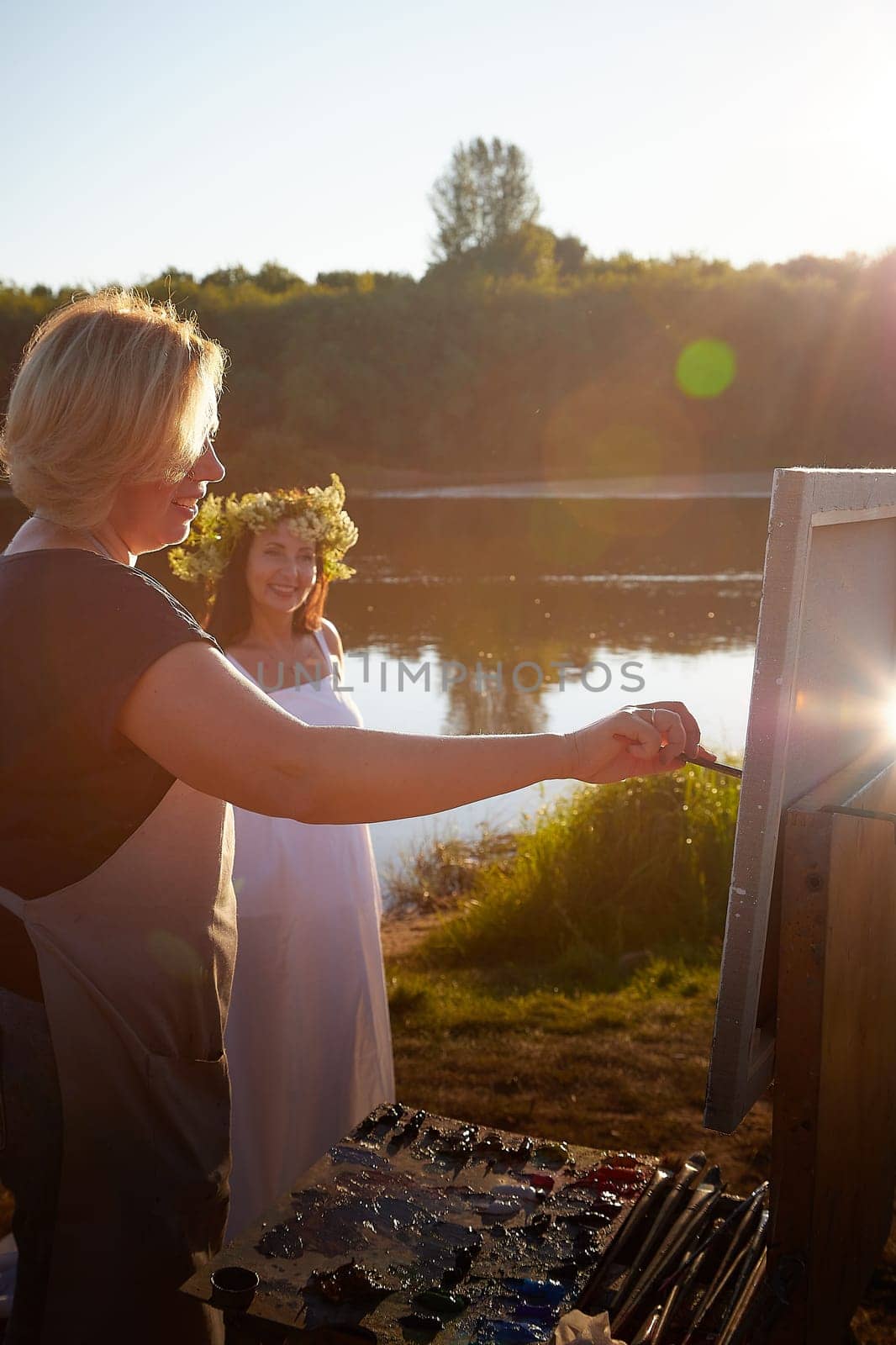 Adult female artist painting picture near water of river or lake in nature and girl in white sundress and flower wreath for the holiday of Ivan Kupala in nature at sunset by keleny