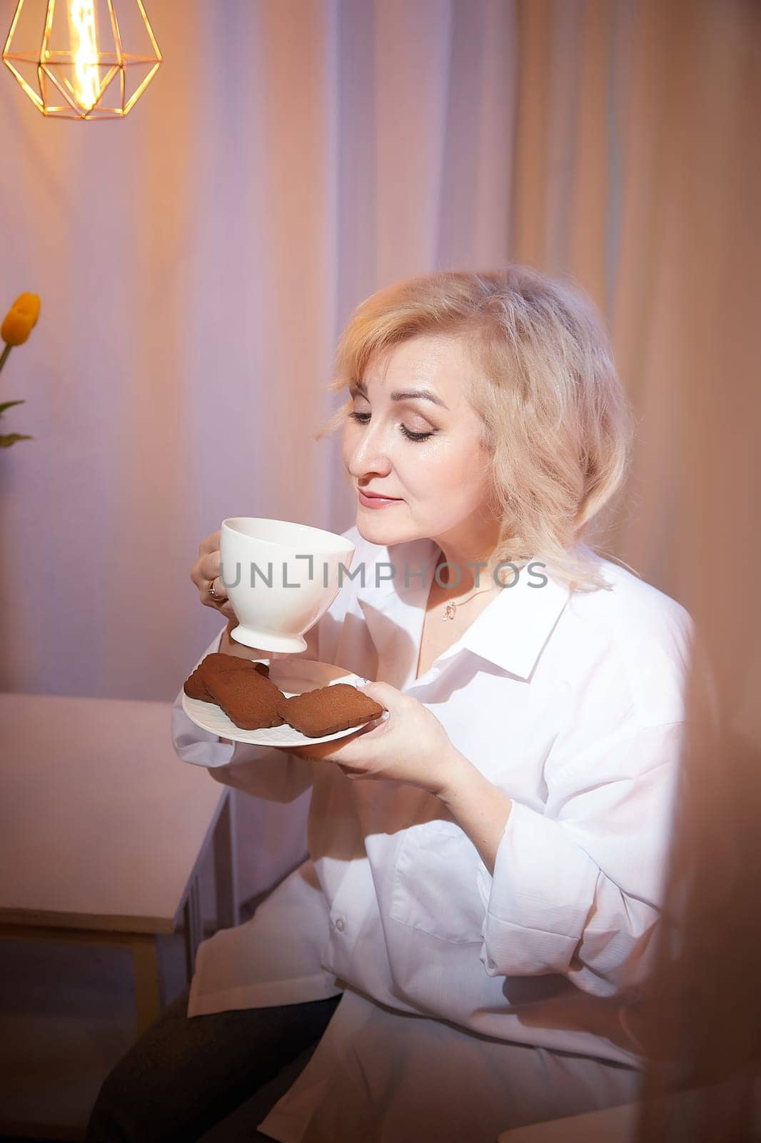 Adult mature woman of 40 or 60 years old with cup of coffee and chocolate cookies in casual white shirt and blue jeans. Calm cozy evening atmosphere with transparent curtains and soft warm lamps by keleny
