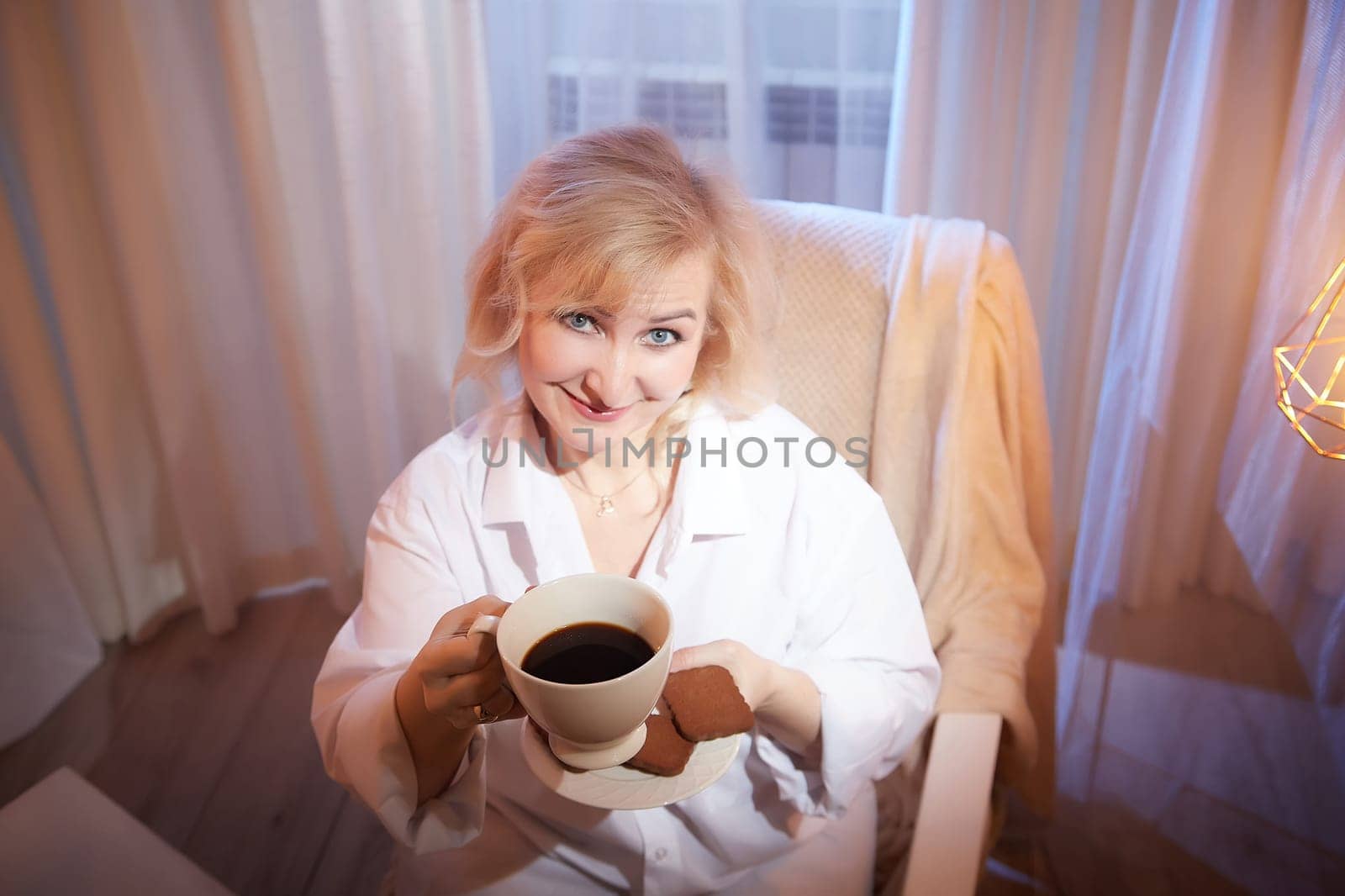 Adult mature woman of 40 or 60 years old with cup of coffee and chocolate cookies in casual white shirt and blue jeans. Calm cozy evening atmosphere with transparent curtains and soft warm lamps by keleny