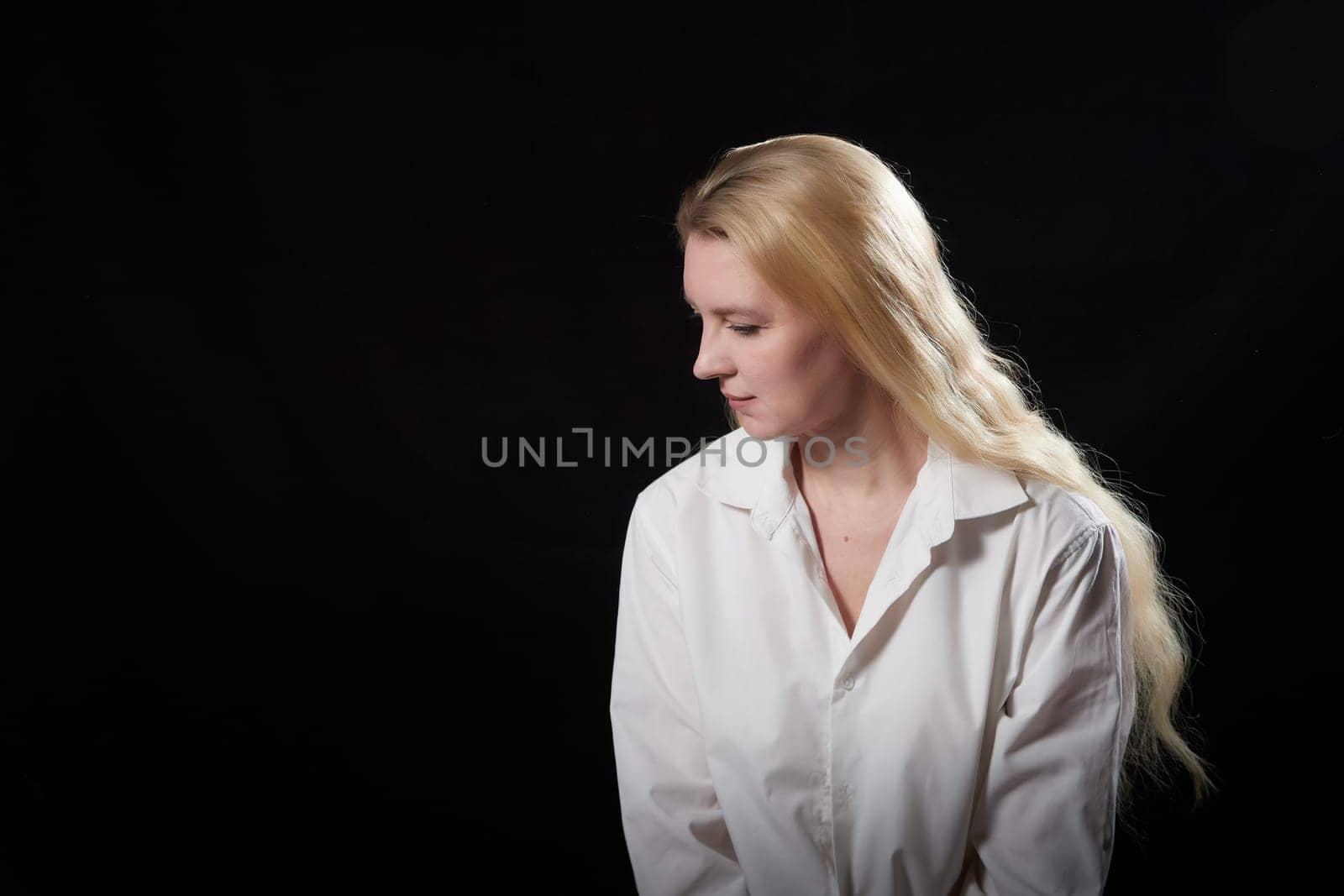 Young beautiful blonde girl in white shirt posing on a black background. Adult woman model posing alone in dark studio