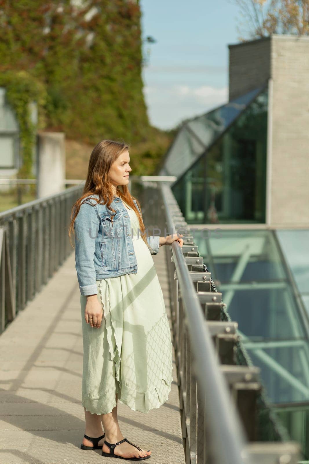 Beautiful white pregnant woman standing near modern eco green high tech building. Young female wears casual clothes. Futuristic eco-friendly estate,green district city architecture Vertical plane