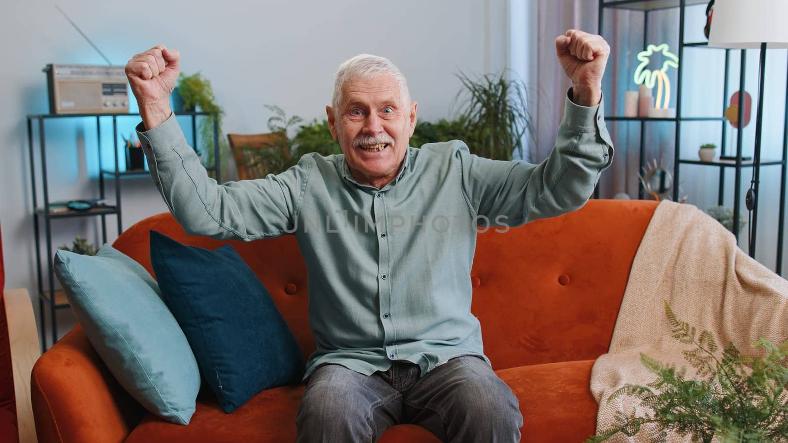 Happy senior grandfather man shouting, celebrating success, winning, goal achievement good news raising fists in gesture I did it, lottery luck at modern home apartment. Elderly guy in room on couch