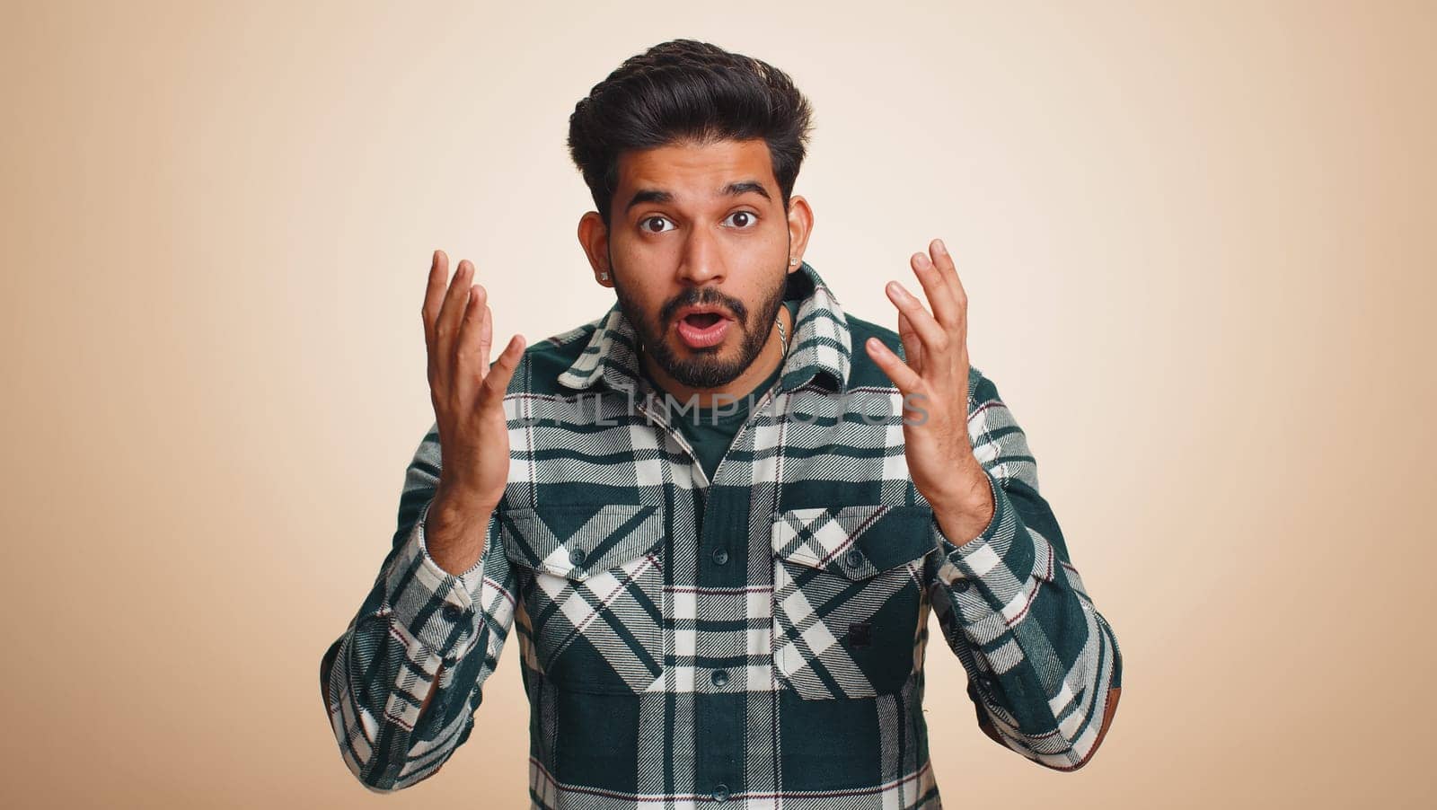 Oh my God, Wow. Excited amazed winner man raising hands in surprise looking at camera with big eyes, shocked by sudden victory. Sincere handsome indian guy isolated alone on beige studio background