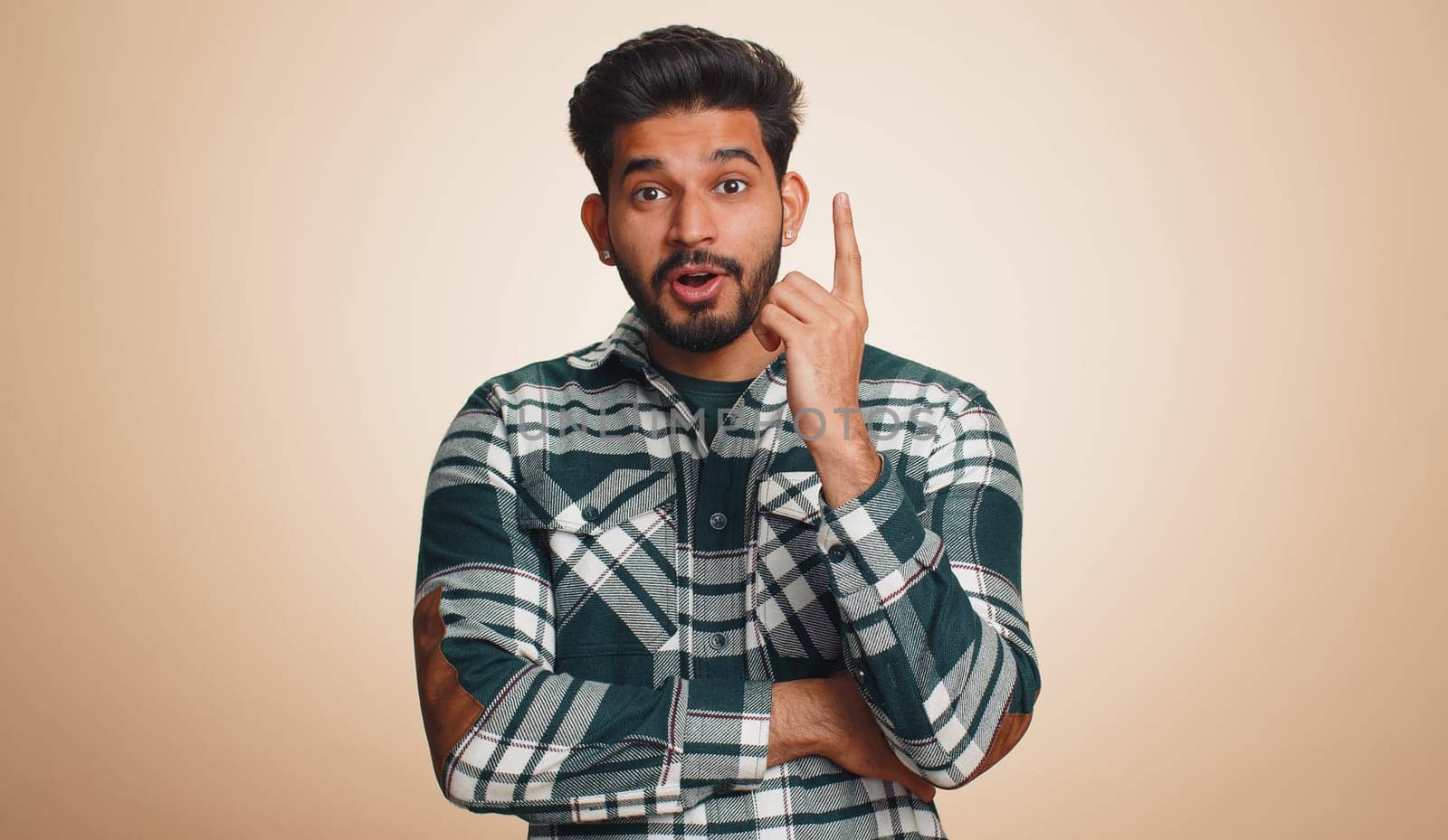 Eureka. Inspired young indian man pointing finger up with open mouth, having good idea, plan, startup, showing inspiration motivation gesture, problem solution. Bearded hindu guy on beige background
