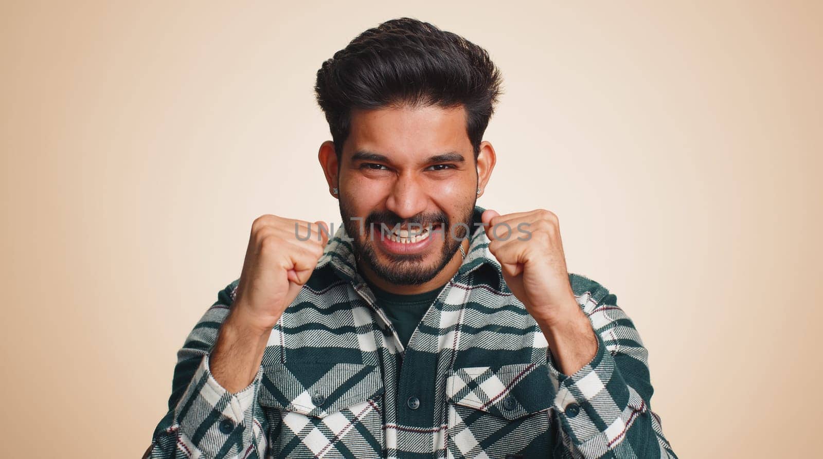 Happy joyful indian man shouting, raising fists in gesture I did it, celebrating success, winning, birthday, lottery jackpot goal achievemen, good news. Young hindu guy on beige studio wall background
