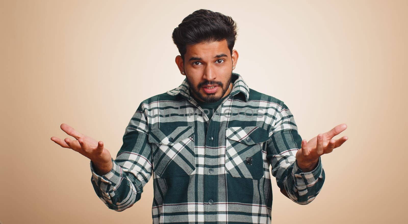 What Why. Sincere irritated indian man raising hands in indignant expression asking reason of failure demonstrating disbelief irritation by trouble. Handsome confused hindu guy on beige background